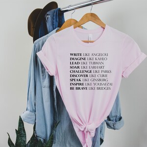 May include: Pink t-shirt with black text that reads "Write like Angelou, Imagine like Kahlo, Lead like Tubman, Soar like Earhart, Challenge like Parks, Discover like Curie, Speak like Ginsburg, Inspire like Yousafzai, Be Brave like Bridges."