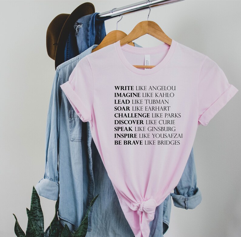 May include: Pink t-shirt with black text that reads "Write like Angelou, Imagine like Kahlo, Lead like Tubman, Soar like Earhart, Challenge like Parks, Discover like Curie, Speak like Ginsburg, Inspire like Yousafzai, Be Brave like Bridges."
