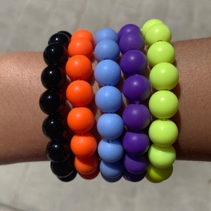 May include: A stack of five colorful bracelets with round beads. The bracelets are black, orange, light blue, purple, and neon green.