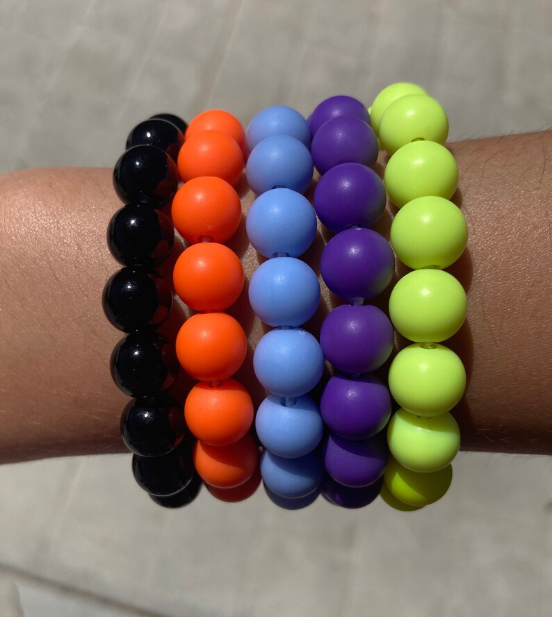 May include: A stack of five colorful bracelets with round beads. The bracelets are black, orange, light blue, purple, and neon green.