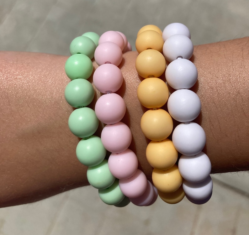 May include: Three colorful beaded bracelets stacked on a wrist. The top bracelet is white, the middle bracelet is yellow, and the bottom bracelet is green.