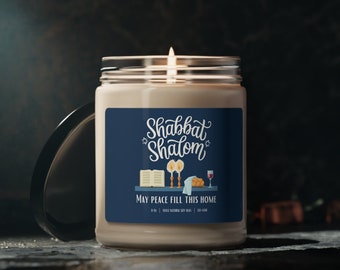 May include: A lit candle in a clear glass jar with a navy blue label. The label reads "Shabbat Shalom" with illustrations of a book, candles, and wine. The text "May Peace Fill This Home" is also on the label. The candle is on a dark surface.