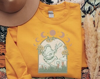 Puede incluir: Una sudadera amarilla con un gráfico de una rana tocando una guitarra rodeada de girasoles y lunas. El gráfico es de estilo vintage.