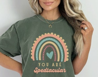 Può includere: T-shirt verde oliva con un design arcobaleno colorato e la scritta "YOU ARE Spedtacular". L'arcobaleno presenta tonalità di rosa, blu e giallo. La maglietta è in stile casual.