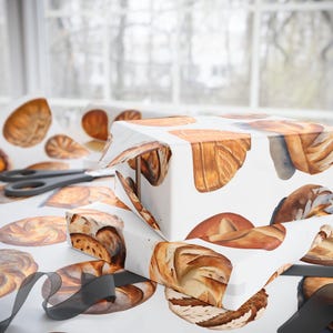 K&ouml;nnte beinhalten: Zwei Geschenkboxen, die in wei&szlig;es Papier mit einem Muster aus Aquarell-Illustrationen verschiedener Brote und Geb&auml;ckst&uuml;cke eingewickelt sind. Die Boxen sind mit schwarzem Band gebunden.