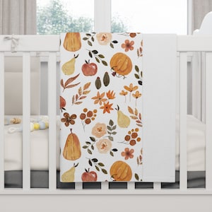 Può includere: Una copertina per neonati bianca con un motivo di zucche arancioni, pere, mele e fiori. La copertina è realizzata in tessuto morbido e confortevole, perfetta per tenere un bambino al caldo e comodo.