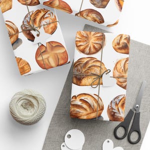 K&ouml;nnte beinhalten: Ein Set aus drei wei&szlig;en Geschenkpapieren mit einem Aquarelldesign aus verschiedenen Broten und Br&ouml;tchen. Die Papiere sind mit Schnur und wei&szlig;en Papieretiketten verziert.