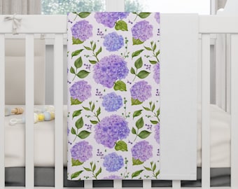 Op de afbeelding: Een witte babydeken met een paars en blauw bloemenpatroon. De deken heeft een herhalend patroon van hortensia's en bladeren.