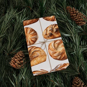 K&ouml;nnte beinhalten: Wei&szlig;es Geschenkpapier mit einer braunen Kordelschleife. Das Geschenkpapier zeigt ein sich wiederholendes Muster aus Aquarell-Illustrationen verschiedener Brotsorten.