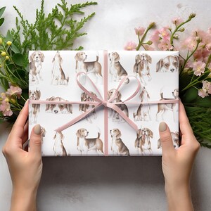 May include: A white gift box with a pink ribbon is wrapped in paper with a repeating pattern of brown and white beagle dogs. The box is surrounded by green foliage and pink flowers.