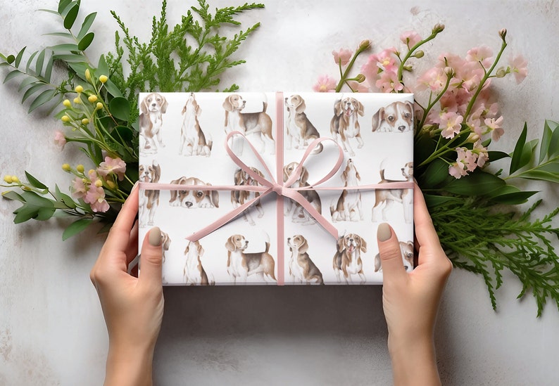 May include: A white gift box with a pink ribbon is wrapped in paper with a repeating pattern of brown and white beagle dogs. The box is surrounded by green foliage and pink flowers.