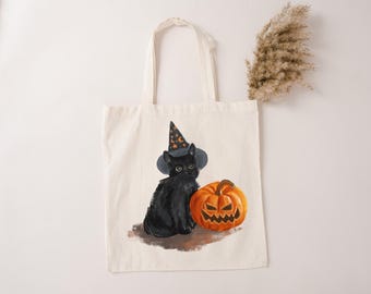 May include: Off-white canvas tote bag featuring a black cat wearing a witch hat and sitting next to an orange jack-o'-lantern. The cat has green eyes. The tote bag has two handles and is suitable for carrying items.