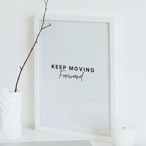 Pode incluir: Impressão emoldurada branca com o texto "KEEP MOVING Forward" em letras pretas. A impressão está sobre uma superfície branca com um vaso branco e uma vela branca.