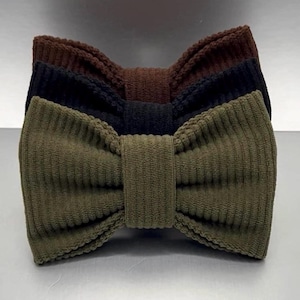 May include: A set of three bow ties made from a textured corduroy fabric. The bow ties are stacked vertically, featuring an olive green one at the front, a black one in the middle, and a dark brown one at the back, each with a central knot.