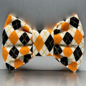 May include: A black, white and orange bow tie with a gold glitter pattern. The bow tie is made of fabric and has a diamond pattern.