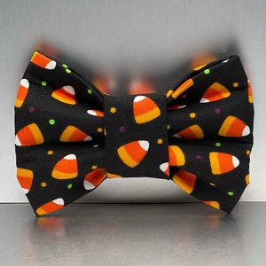 May include: A black bow tie with a candy corn pattern. The bow tie has orange, white, and yellow candy corn on a black background with green and purple polka dots.