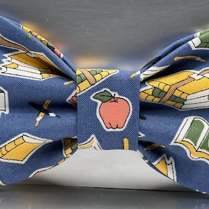 May include: A blue bow tie with a repeating pattern of books, apples, and pencils. The bow tie features a central apple design and is suitable for a teacher or student.