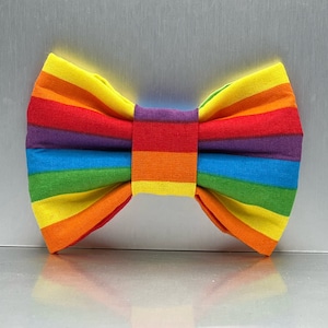 May include: A rainbow striped bow tie made of fabric. The bow tie is yellow, orange, red, purple, blue, and green.