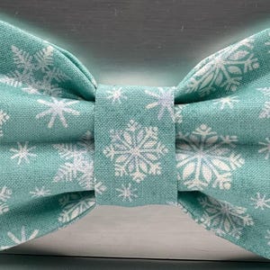 May include: A teal bow tie featuring a white snowflake pattern. The fabric bow tie is tied in a classic style, with a central band. The snowflakes are of varying sizes and designs, creating a winter theme.