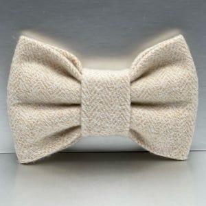May include: A beige bow tie with a textured, woven appearance. The bow tie is a neutral color, with a classic bow shape. The fabric has a subtle herringbone pattern.