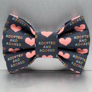 May include: A dark gray fabric bow tie featuring a repeating pattern of light pink hearts and the text ADOPTED AND ADORED printed in alternating blue, yellow, and orange letters across the surface of the bow tie.