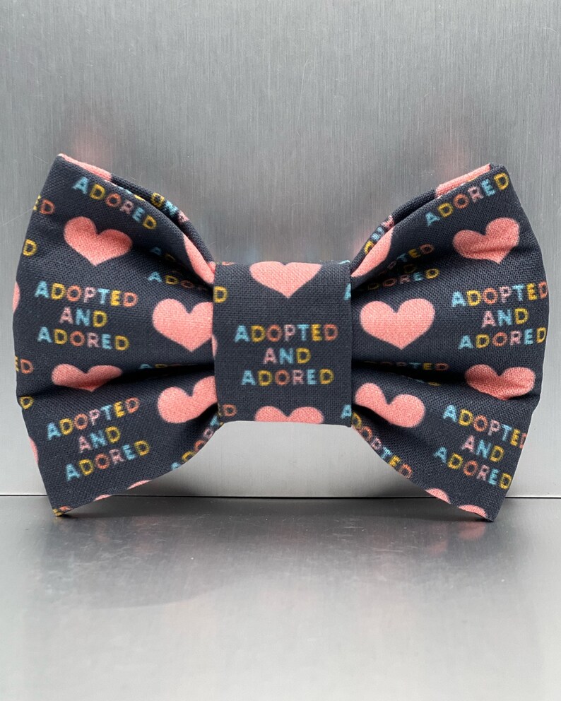 May include: A dark gray fabric bow tie featuring a repeating pattern of light pink hearts and the text ADOPTED AND ADORED printed in alternating blue, yellow, and orange letters across the surface of the bow tie.
