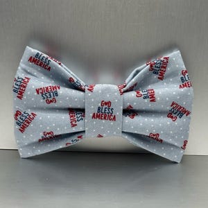 Peut inclure: Un nœud papillon bleu clair avec un motif à pois blancs. Le nœud papillon porte les mots "GOD BLESS AMERICA" imprimés en rouge et bleu. Le nœud papillon est en tissu et est probablement fait à la main.