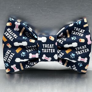 May include: A dark navy blue bow tie featuring a repeating pattern of white dog bones, light blue paw prints, and tan treats. The text "TREAT TASTER" is printed in white across the fabric, including on the central knot of the bow tie.