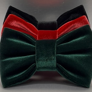 May include: A velvet bow tie in a layered design of black, red, and green. The bow tie has a classic shape with a central knot and two symmetrical loops. The fabric has a soft, plush texture, and the colors are rich and saturated.
