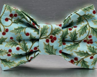 May include: A light blue bow tie featuring a repeating pattern of green holly leaves and red berries. The leaves are outlined in a thin gold metallic thread, creating a festive botanical design across the fabric surface of the accessory.
