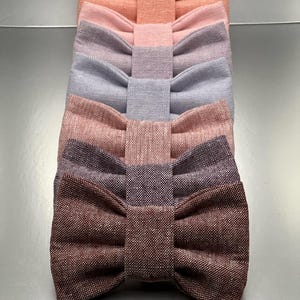 May include: A vertical stack of six bow ties made from a textured fabric. The colors range from a light peach at the top, followed by soft pink, light lavender, periwinkle blue, dusty rose, and a deep dark brown at the bottom of the stack.