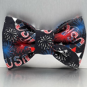 May include: A bow tie with a black background, decorated with red, white, and blue fireworks and the letters "USA". This bow tie is a festive accessory, suitable for patriotic events.