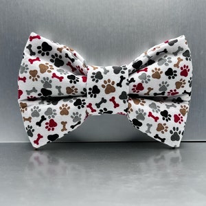 May include: White bow tie with a paw print and bone pattern in shades of brown, gray, red, and black.