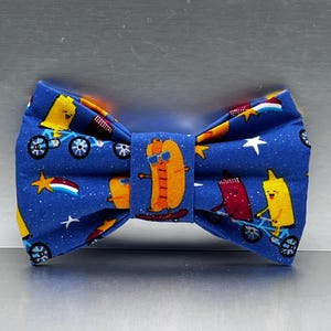 May include: A blue bow tie with a repeating pattern of cartoon hot dogs, mustard, and ketchup riding bicycles and skateboards, with white stars and shooting stars. The bow tie is made of fabric.
