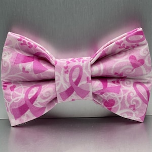 May include: A fabric bow tie featuring a repeating pattern of pink awareness ribbons, small pink hearts, and white swirling flourishes on a light pink background. The bow tie has a structured, pleated center knot and a classic double-wing shape.