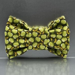 May include: A bow tie featuring a repeating pattern of light green apples with small brown stems and leaves set against a dark brown background. The fabric has a visible woven texture, and the bow tie is constructed with a central knot.