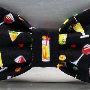 May include: A black bow tie with a repeating pattern of colorful cocktail glasses, cherries, and citrus fruits. The bow tie is a classic shape, suitable for formal or semi-formal occasions. The fabric has a smooth texture.