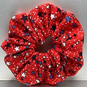 May include: Red scrunchie featuring a pattern of white dots and navy blue and white stars. This hair accessory is a fun and festive addition to any hairstyle. The scrunchie is made of a soft fabric.
