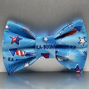Peut inclure: Un nœud papillon bleu avec un motif de feux d'artifice patriotique. Le nœud papillon présente des fusées, des étoiles et des éclats rouges, blancs et bleus sur un fond bleu clair. Le texte "KA-BOOM" est imprimé au centre.