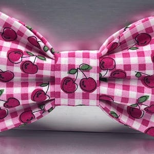 May include: A pre-tied bow tie featuring a pink and white gingham checkered pattern. The fabric is decorated with a repeating print of small red cherries with green stems and leaves, arranged in a symmetrical, pleated bow tie construction.