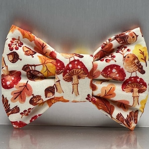 May include: A cream-coloured bow tie with a fall-themed pattern. The fabric is printed with illustrations of red mushrooms, orange and yellow leaves, acorns, and small brown birds. The bow tie is tied in a classic shape.