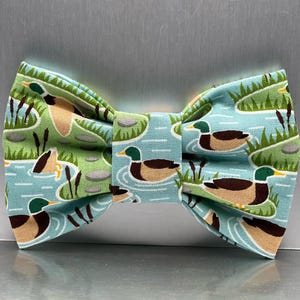 May include: A bow tie with a duck pond pattern. The bow tie features a light blue background with illustrations of ducks, reeds, and water. The ducks have brown bodies, green heads, and yellow beaks. The bow tie is a classic accessory.