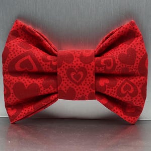 May include: A red fabric bow tie featuring a repeating pattern of dark red hearts and a subtle dotted texture. The bow tie has a structured, pleated design with a central fabric band that holds the folds in place, creating a symmetrical shape.