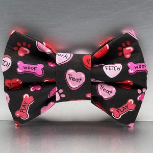 May include: A black bow tie featuring a repeating pattern of pink and red hearts and bones. The hearts are printed with the words Treat, Bark, and Paw, while the bones feature the words Woof, Fetch, and Play, along with small paw print icons.