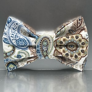 May include: A pre-tied bow tie featuring a white base fabric with a detailed paisley and floral pattern in shades of blue, brown, and muted green. The fabric has a visible woven texture, and the design is symmetrical with a central knot detail.