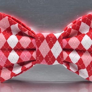 May include: A red and white argyle patterned bow tie. The bow tie features a repeating diamond pattern in red, white, and pink. The bow tie is made of fabric and is tied in the center.