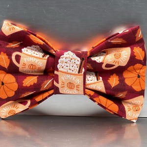May include: A burgundy bow tie featuring a repeating pattern of orange pumpkins, autumn leaves, and pumpkin spice latte mugs. The mugs have the text "Pumpkin Lover". A seasonal accessory for the autumn season.