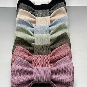May include: A collection of eight bow ties arranged in a vertical stack, featuring a variety of solid colors including white, black, charcoal, beige, light blue, sage green, olive green, red, and mauve, all crafted from a textured fabric material.