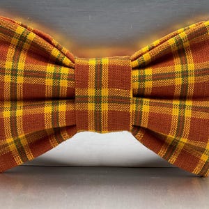 May include: A plaid bow tie in shades of brown, yellow, and green. The bow tie features a classic plaid pattern, with a symmetrical design. The fabric appears to be woven, giving it a textured look. The bow tie is a fashion accessory.