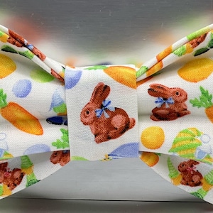 May include: A white bow tie featuring a repeating pattern of brown bunnies with blue bows, orange carrots with green tops, yellow and blue Easter eggs, and green and yellow striped gnome hats. The fabric has a textured, woven appearance.